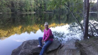 A woman in a pink sweater sits on a large stone next to a lake or river in a picturesque, wooded, nature scene.