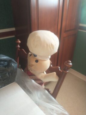 A small, stuffed Pillsbury Doughboy is seated on a chair at the author's dining room table. The walls are a deep green, and there's a large wooden cabinet in the background.